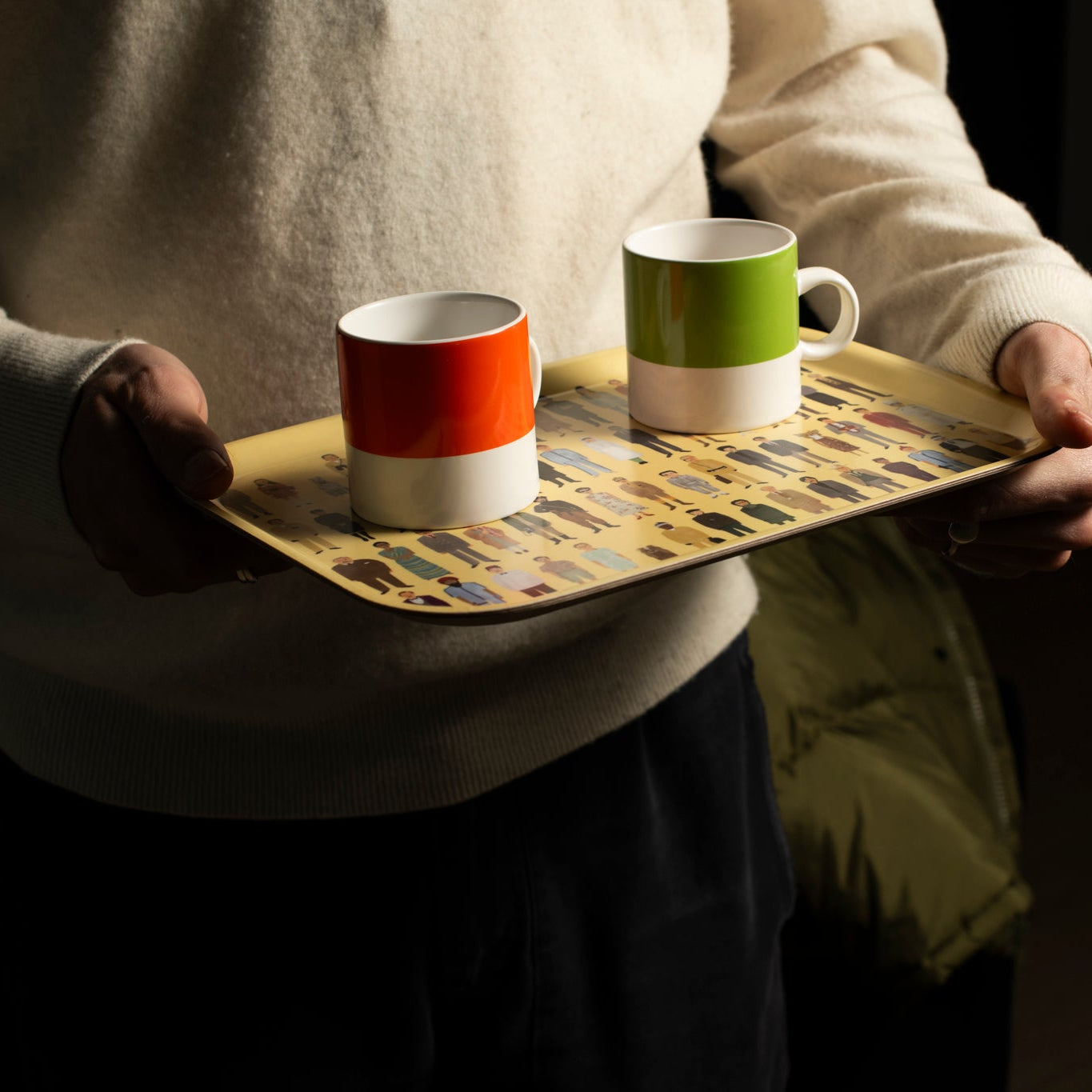 Person holding a yellow tray with two colorful mugs against a dark background