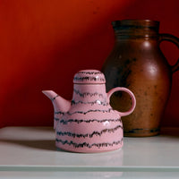 A pink and black abstract ceramic coffee pot on a white surface against an orange wall.