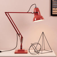 A red adjustable desk lamp with a rectangular base and articulated arm, shown lit on a white tabletop against a pale pink wall. A black wire geometric sculpture and a framed picture are also on the table.