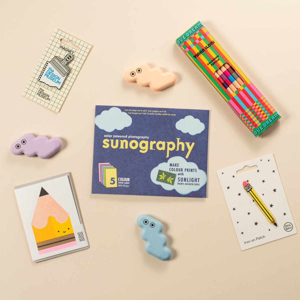 A styled flat-lay of stationery items on a warm beige background, including Sunography cards, colouring pencils, a pencil card, and three pastel erasers.