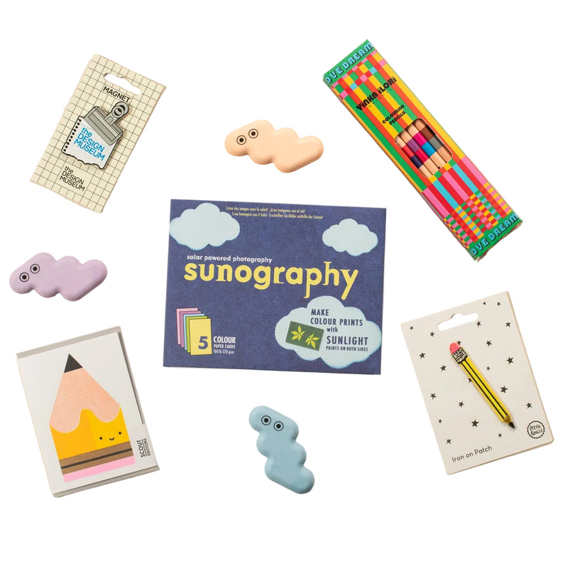 A styled flat-lay of stationery items on a warm beige background, including Sunography cards, colouring pencils, a pencil card, and three pastel erasers.