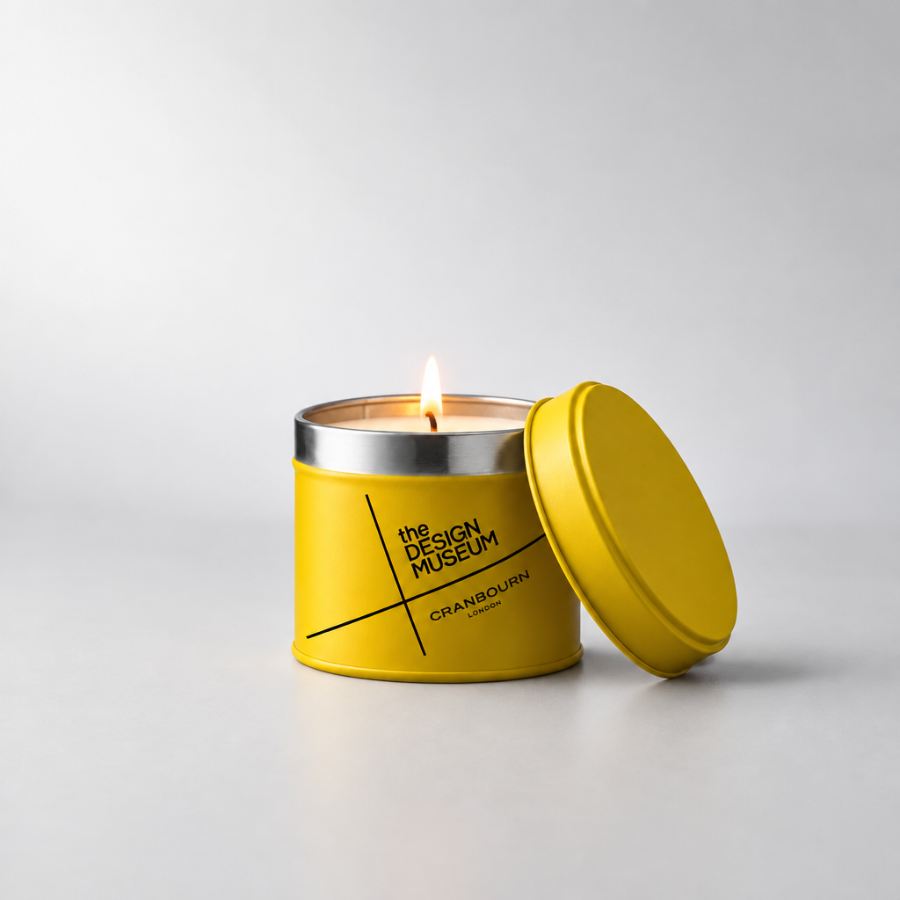 A yellow metal candle tin with its lid leaning against it on a light gray studio background. The tin has a silver rim and visible wick, with black printed text reading 'the DESIGN MUSEUM' and 'GRANBOURN'.