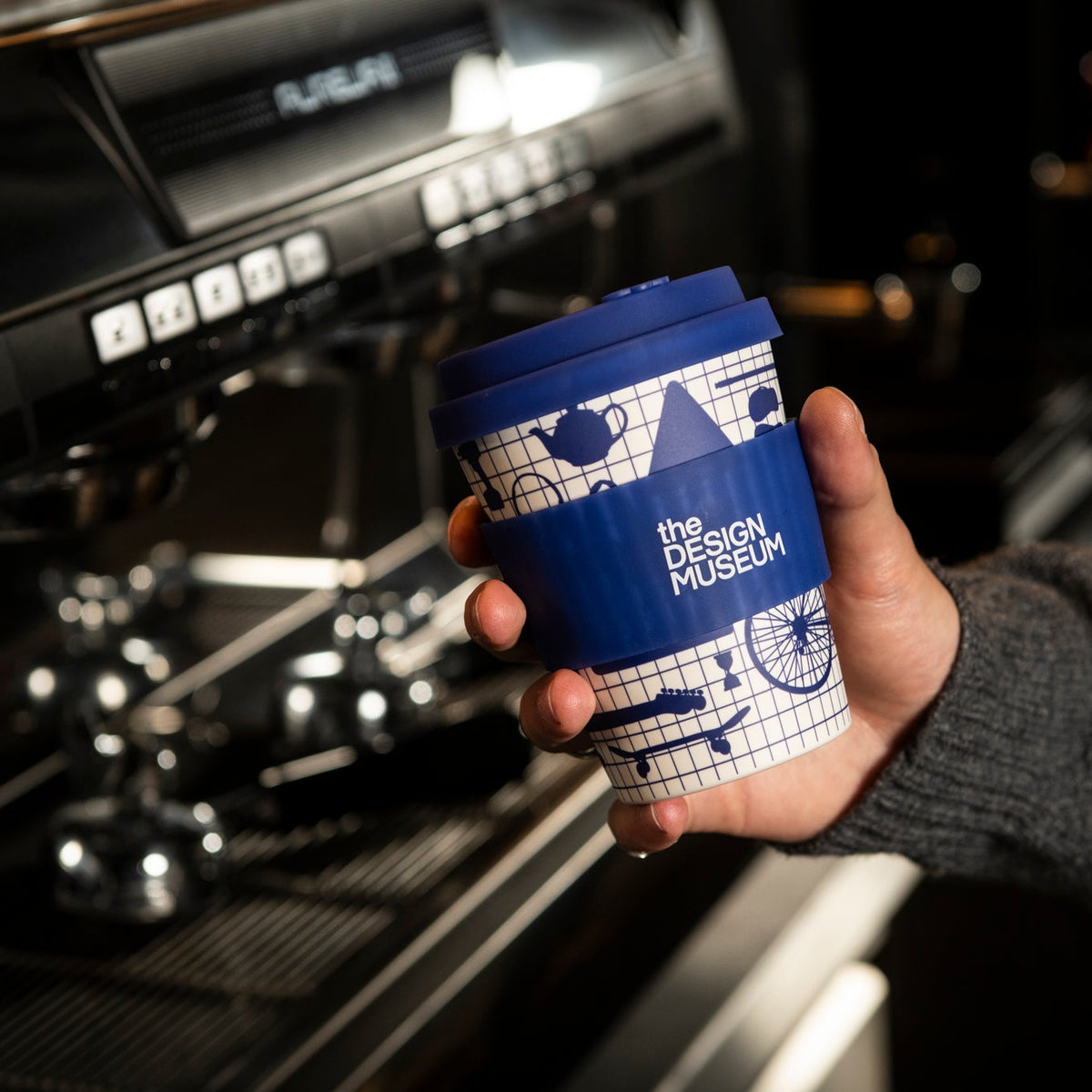 Hand holding a blue and white coffee cup with a design museum logo in front of a coffee machine.