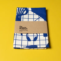 A folded white tea towel with blue designs of everyday objects with a cardboard belly band on a yellow background.