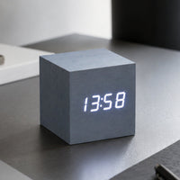 A cube-shaped digital clock with a matte gray concrete-like finish and glowing white LED numerals, on a grey desk 