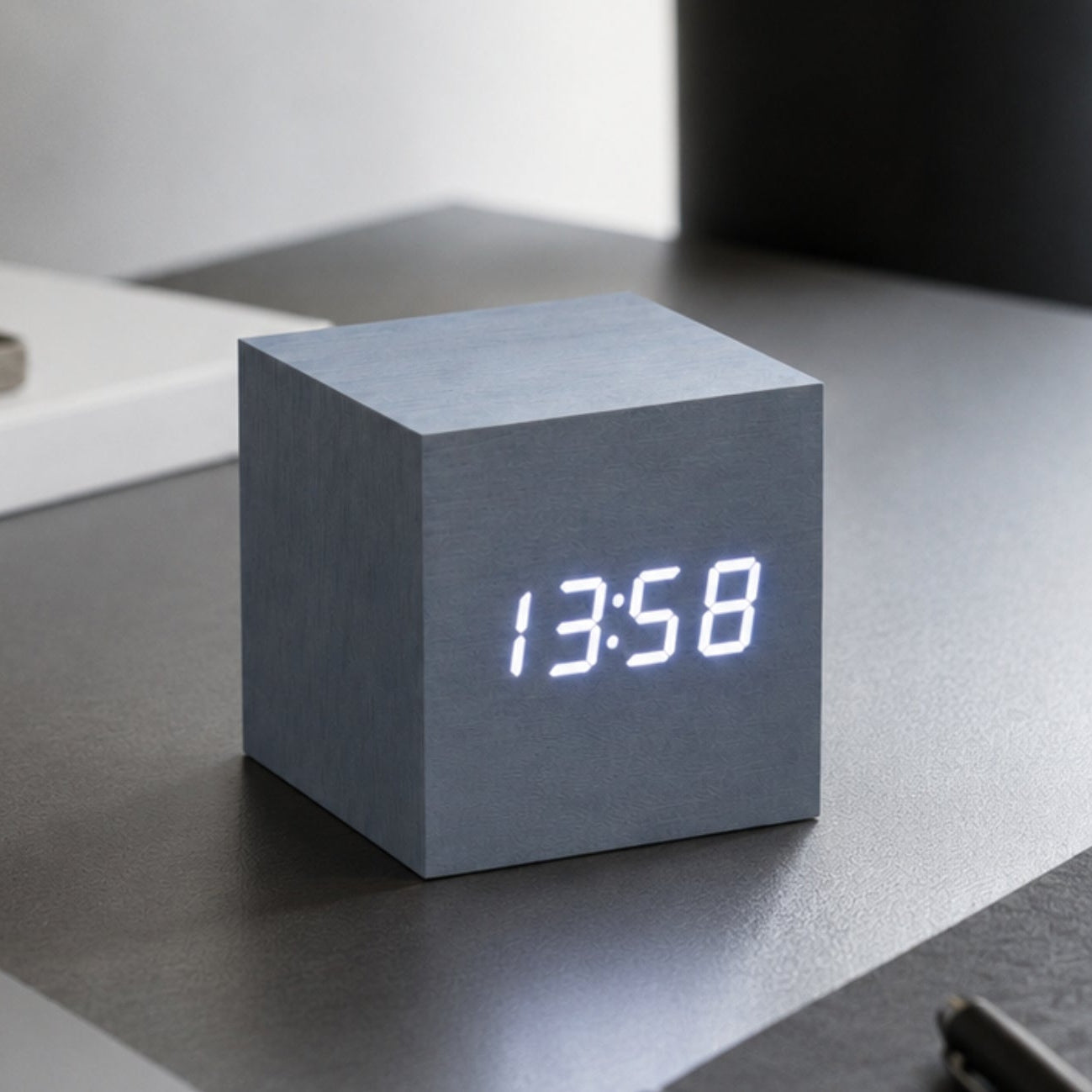 A cube-shaped digital clock with a matte gray concrete-like finish and glowing white LED numerals, on a grey desk 