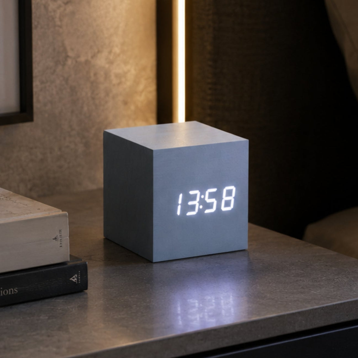 A modern cube-shaped digital clock with a matte gray finish and glowing white LED numbers, on a dark wooden bedside table.