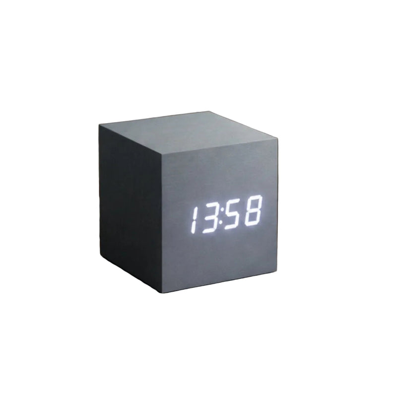 A cube-shaped digital clock with a matte light blue-gray finish sits on a rustic wooden nightstand, displaying the time in glowing white LED digits.