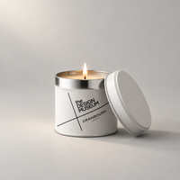A white metal candle tin with a silver rim and a lit wick, shown on a light gray studio background with the lid leaning beside it. The front label reads 'the DESIGN MUSEUM' and 'CRANBOURN LONDON'.