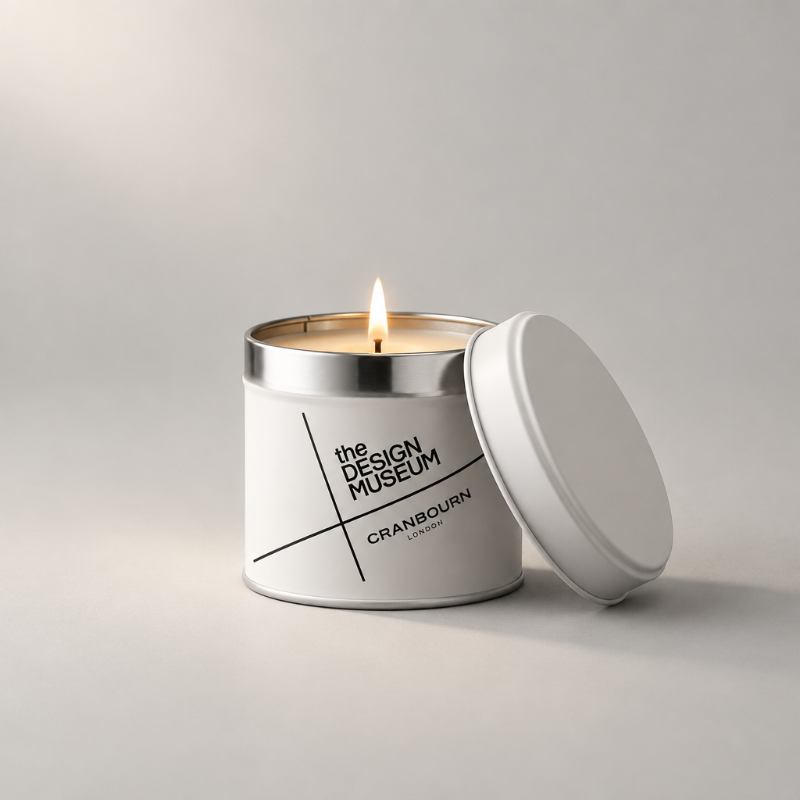 A white metal candle tin with a silver rim and a lit wick, shown on a light gray studio background with the lid leaning beside it. The front label reads 'the DESIGN MUSEUM' and 'CRANBOURN LONDON'.