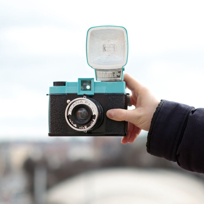 A hand holding up a blue and black vintage camera with a flash attachment against a blurred background.