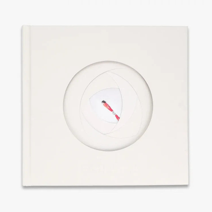 A square white book  with a circular cutout in the center, showing a small red and black abstract mark inside. The item is photographed against a clean white background.