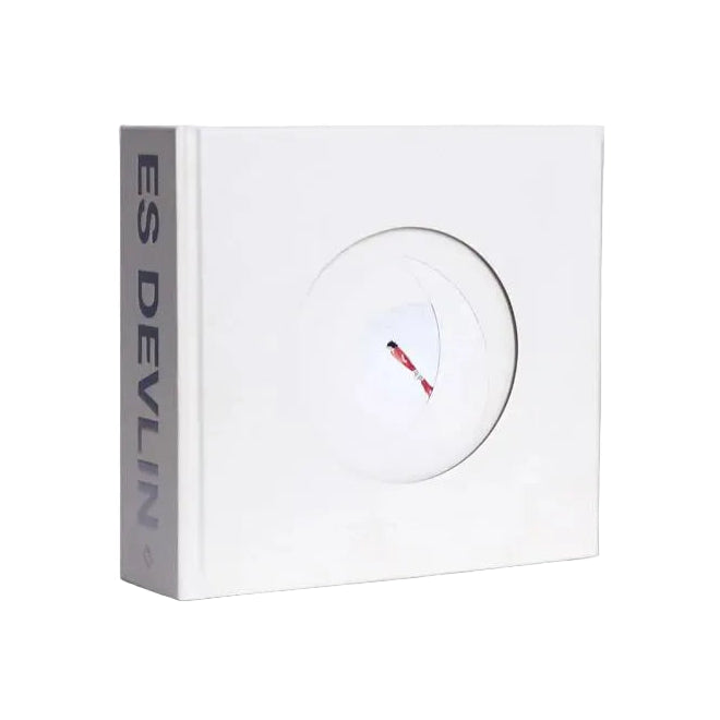 A white square book with a circular cutout on the front, showing a small red and black object inside, and the vertical text 'ES DEVILIN' printed on the side panel, set against a plain white background.