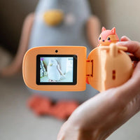 Orange kids camera with screen held by a hand, with a lens showing a blurred background.