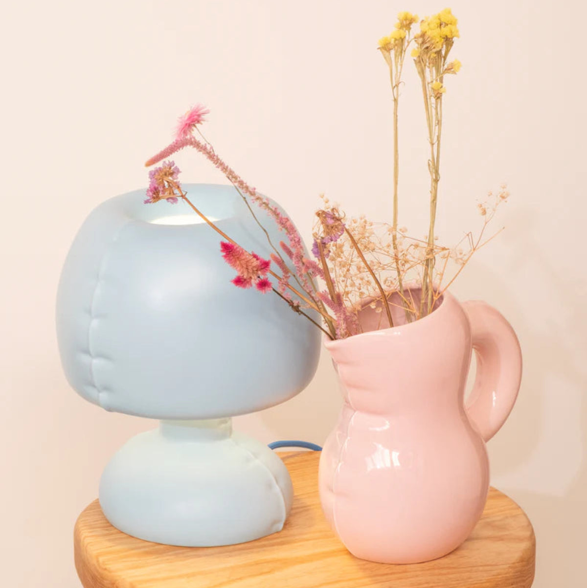 Light blue lamp and pink pitcher with dried flowers on a wooden stool against a beige background