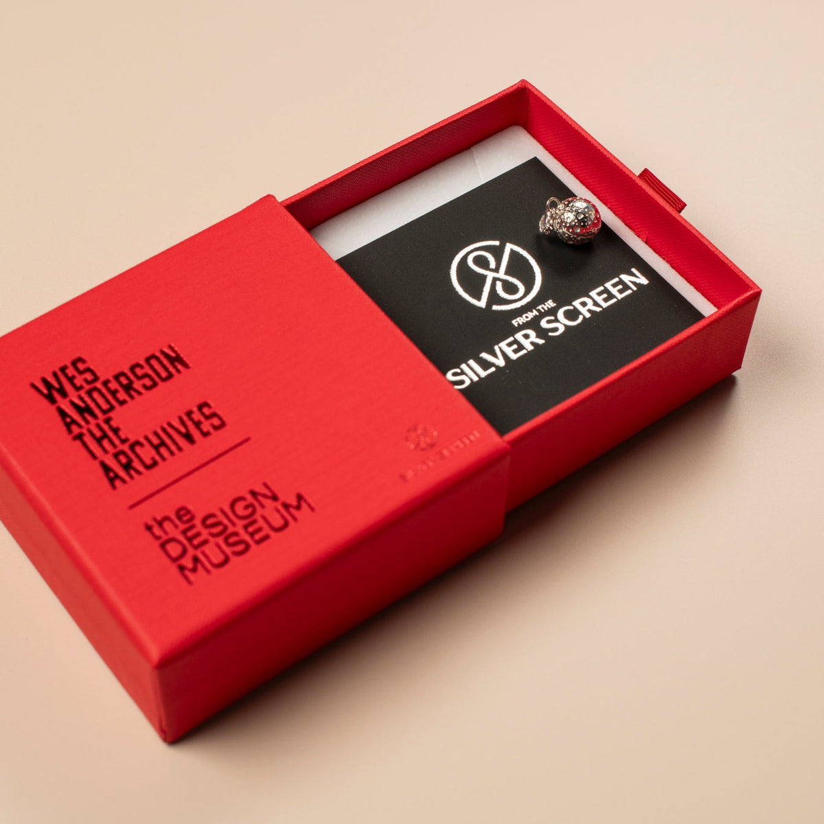 Red jewellery box with a black card and a silver charm featuring 'The Design Museum' logo on a beige background