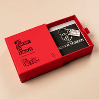 Red jewellery box with a silver clapperboard keychain inside on a beige background 