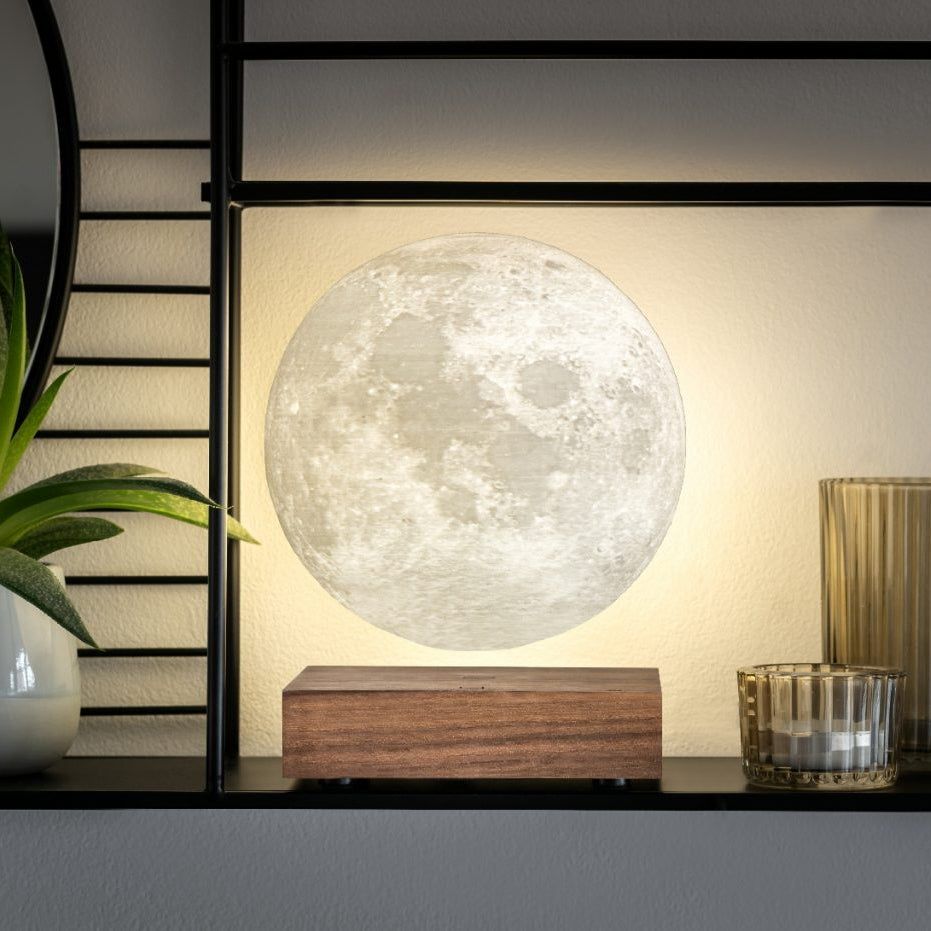 A glowing moon lamp on a wooden base displayed on a shelf next to a potted plant and decorative glassware.