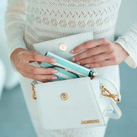 White handbag with gold accents and a blue and white striped glasses case held by a person, against a light blue background