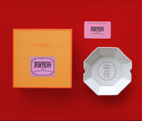White octagonal ceramic ashtray on a red background, next to an orange box and pink card.
