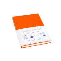 An orange hardcover notebook or guidebook with a white paper band around the middle reading ' grids & guides' in purple text, photographed on a clean white background.