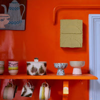 A collection of colourful abstract ceramic bowls and mugs on an orange shelf.
