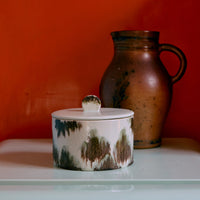 A green and black abstract ceramic storage jar on a white surface in front of an orange wall.