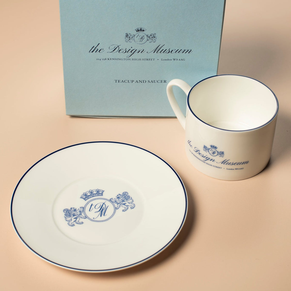 White ceramic cup and saucer with 'The Design Museum' branding next to a blue box on a beige surface.