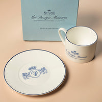 White ceramic cup and saucer with 'The Design Museum' branding next to a blue box on a beige surface.
