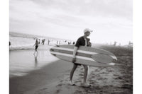 Black and white vintage photo of a person carrying a surfboard on a beach 
