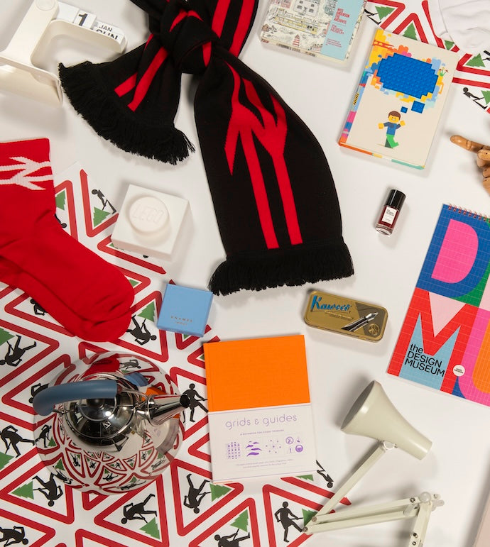 A styled flat-lay of a black knit scarf with red graphic stripes and black fringe, surrounded by various colorful objects on a white surface with a red and green geometric patterned background.