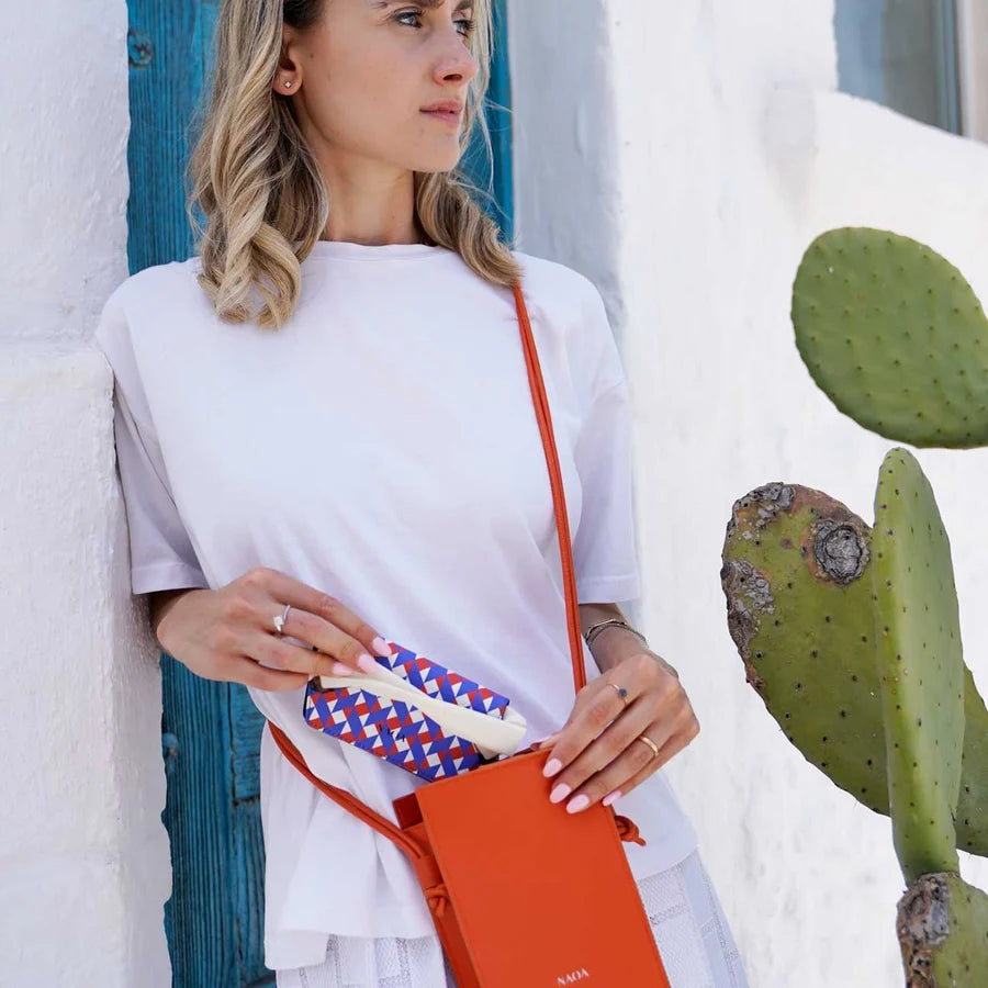 A holding a bright orange bag and a patterned glasses case against a white wall next to a cactus.