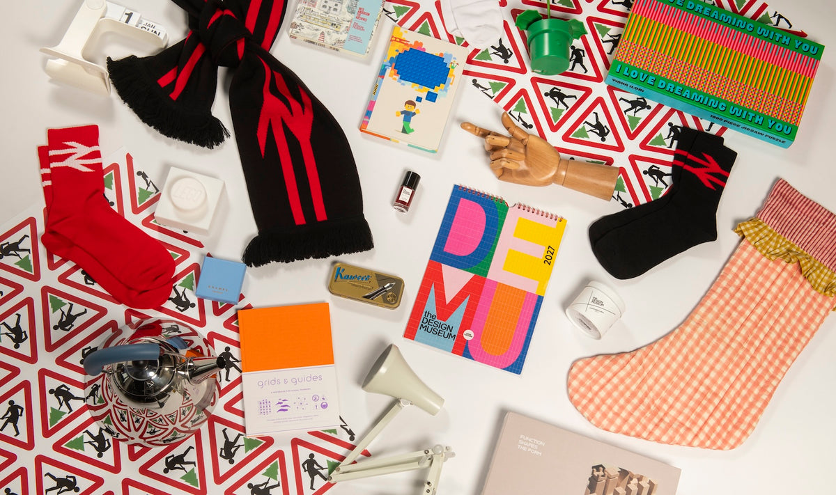 A styled flat-lay of multiple colorful items on a white surface with a red-and-white geometric patterned fabric underneath. Visible items include a black scarf with red stripes, red socks, a black sock, an orange-and-white checkered stocking, a patterned notebook, a small book or card, a notebook with large multicolor letters, a metal teapot, a small lamp, and other assorted objects.