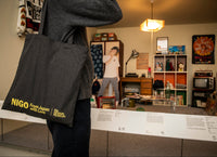 Person holding a black tote bag with 'NIGO' branding in an indoor setting.