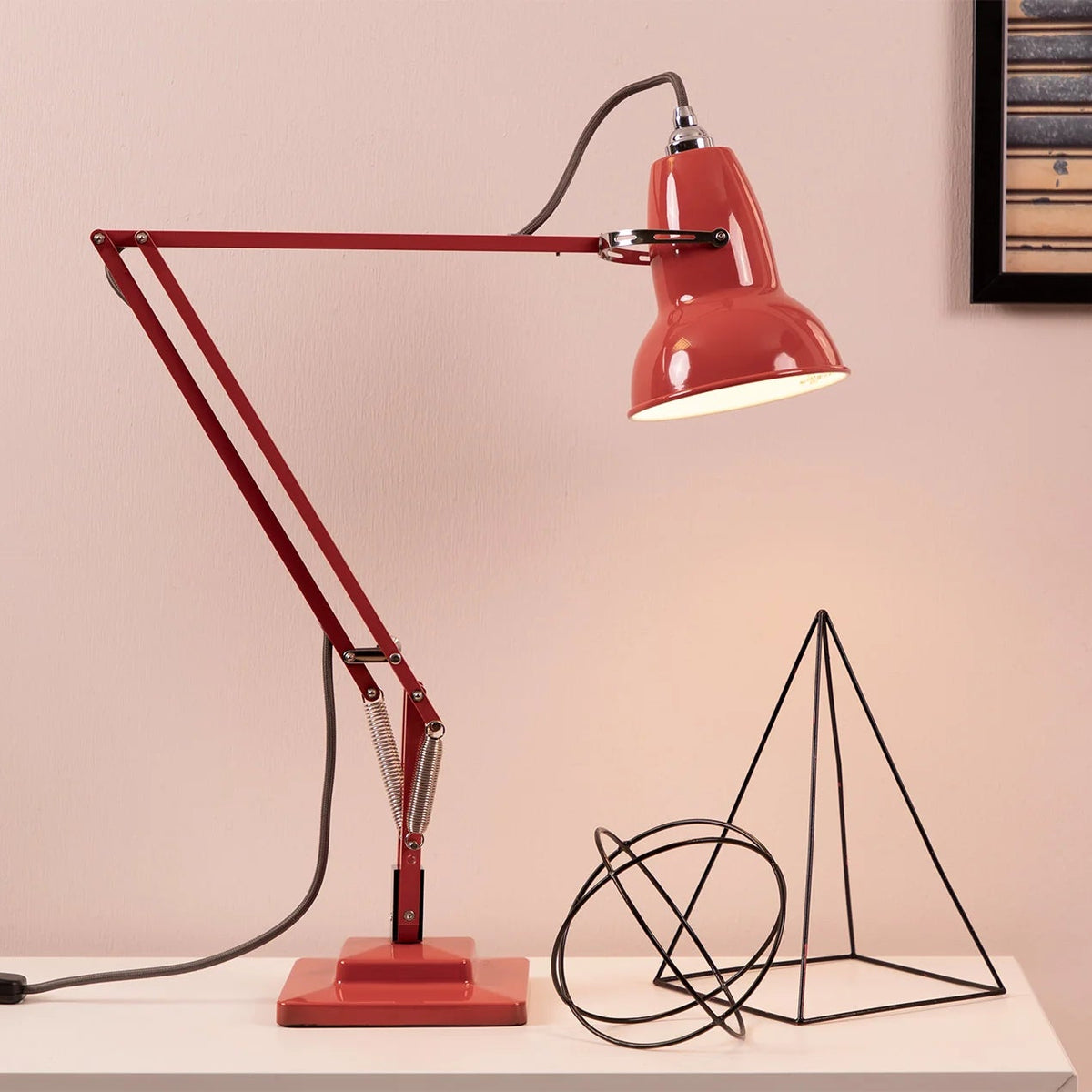 A red adjustable desk lamp with a rectangular base and articulated arm, shown lit on a white tabletop against a pale pink wall. A black wire geometric sculpture and a framed picture are also on the table.