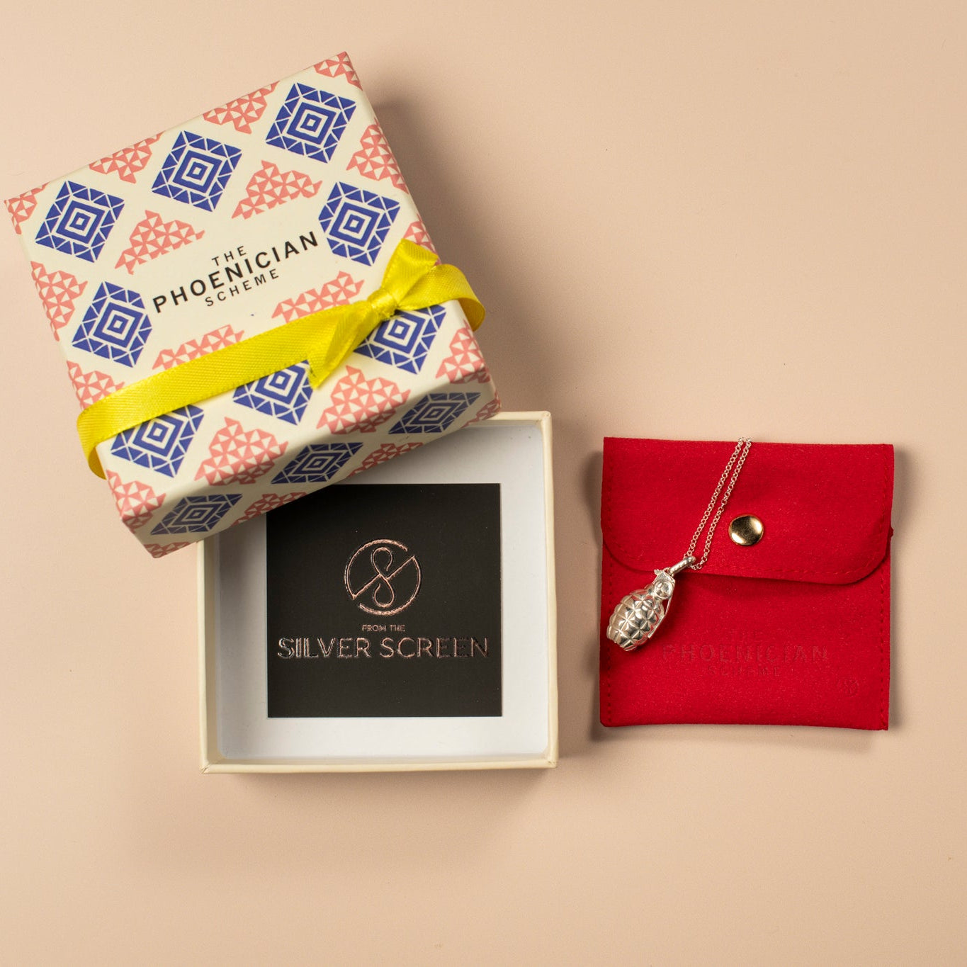Decorative jewellery box with a yellow ribbon, and a silver grenade necklace on a red pouch against  a beige background
