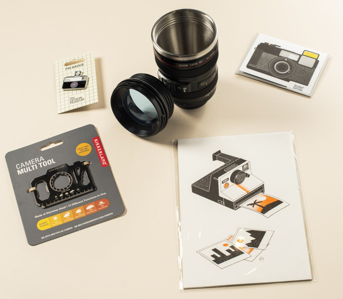 A camera-shaped travel mug on a light beige background, alongside a camera multi-tool, a camera print, and a small card.