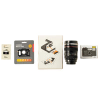 A flat-lay arrangement of camera-related items including a camera multi-tool, a camera print, a black camera lens mug, and a camera pin badge.