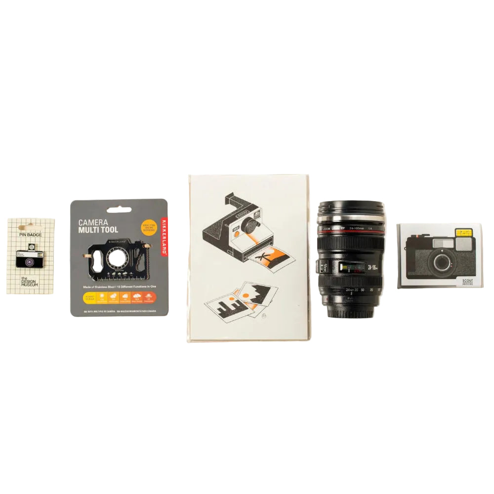 A flat-lay arrangement of camera-related items including a camera multi-tool, a camera print, a black camera lens mug, and a camera pin badge.