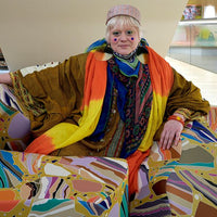 Person wearing a colorful outfit with a rainbow scarf, sitting on a patterned chair.