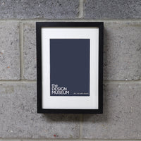 A black picture frame with a white mount, displaying a print titled 'the DESIGN MUSEUM', hung on a grey brick wall.