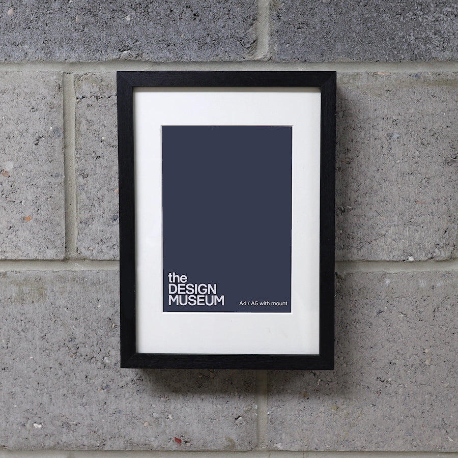 A black picture frame with a white mount, displaying a print titled 'the DESIGN MUSEUM', hung on a grey brick wall.