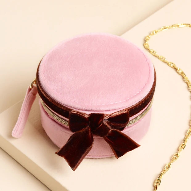 Pink round jewelry box with a brown velvet bow on a beige surface.