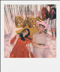 A polaroid picture of two people sitting on a bed with 'BABY' balloons and decorative pillows.