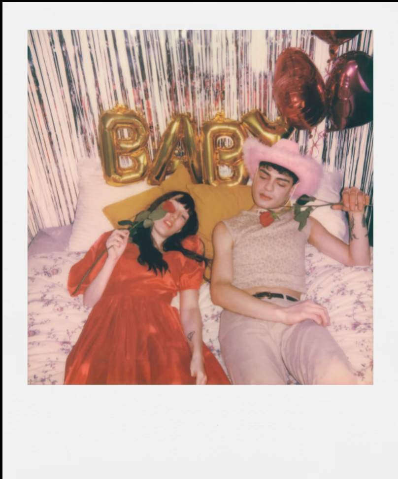 A polaroid picture of two people sitting on a bed with 'BABY' balloons and decorative pillows.