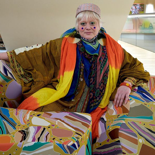 Person wearing a colorful outfit with a multicolored scarf, sitting on a vibrant, abstract-patterned chair.