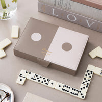 A set of dominoes, shown in a styled flat-lay with several white domino tiles arranged around it on a light gray surface. The box has a split taupe and white design with a circular dot motif 