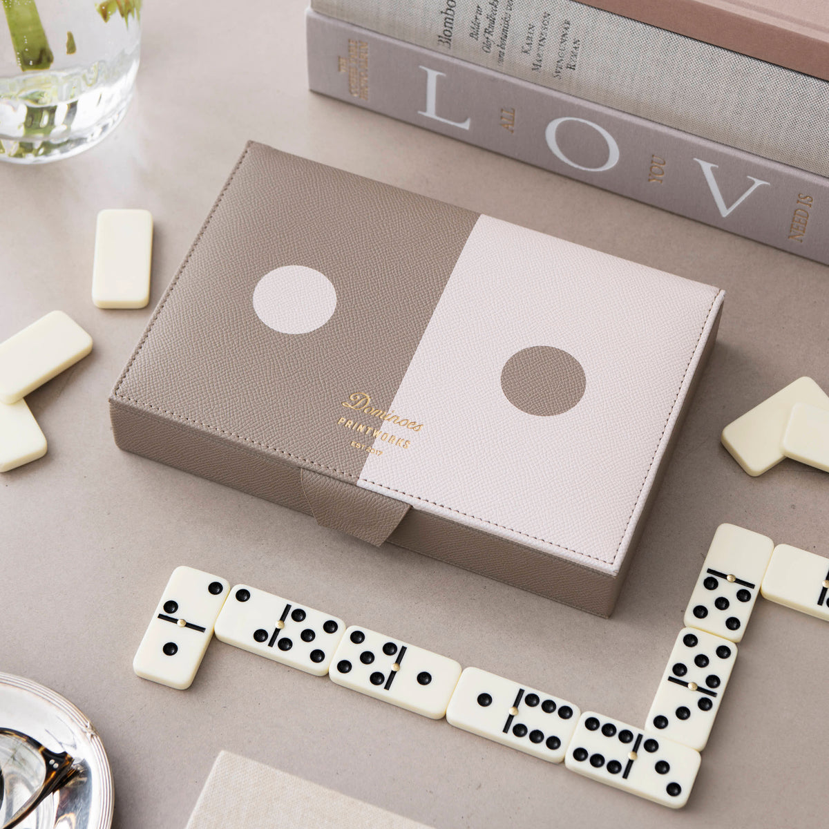 A set of dominoes, shown in a styled flat-lay with several white domino tiles arranged around it on a light gray surface. The box has a split taupe and white design with a circular dot motif 