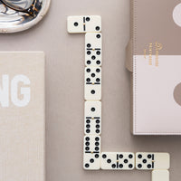 A styled flat-lay featuring a set of cream-colored domino tiles arranged in a domino chain on a neutral gray surface, alongside a textured beige case