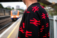 A person standing on a train station platform wearing a black knit sweater with a repeating red railway line logo pattern, with an orange train blurred in the background.